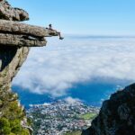 A person sits on a cliff edge enjoying a breathtaking view of the ocean and city below.